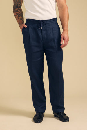 A person wears Velvet by Graham & Spencer’s ELLIOTT LINEN PANT in navy, styled with a white short-sleeve shirt and black shoes, standing against a beige background.