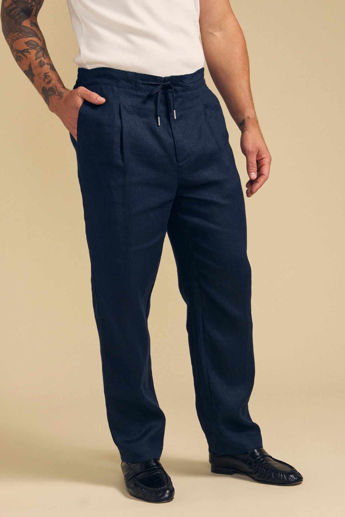 A man wearing a white short-sleeve shirt, Velvet by Graham & Spencer ELLIOTT LINEN PANT in navy, and black loafers stands against a beige background.-41701921751233