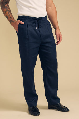 A man wearing a white short-sleeve shirt, Velvet by Graham & Spencer ELLIOTT LINEN PANT in navy, and black loafers stands against a beige background.