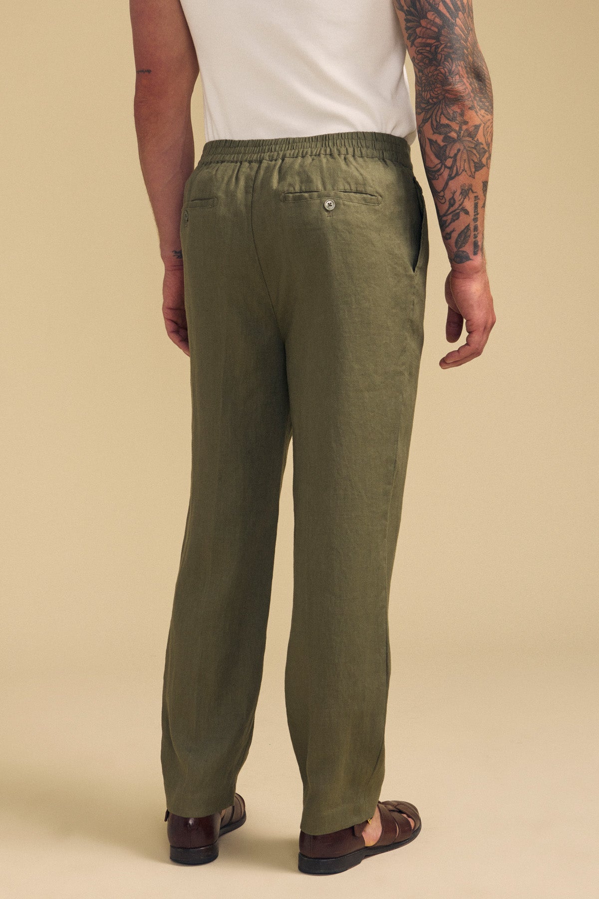 A man wearing Velvet by Graham & Spencer's ELLIOTT LINEN PANT in olive green, paired with a white shirt and brown sandals, stands facing away to display the back of the trousers and his arm tattoos.-41701921587393