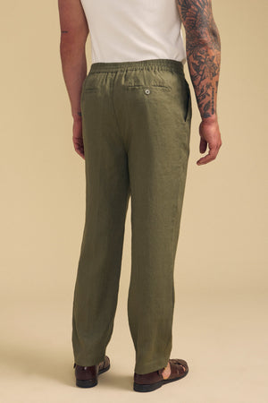 A man wearing Velvet by Graham & Spencer's ELLIOTT LINEN PANT in olive green, paired with a white shirt and brown sandals, stands facing away to display the back of the trousers and his arm tattoos.