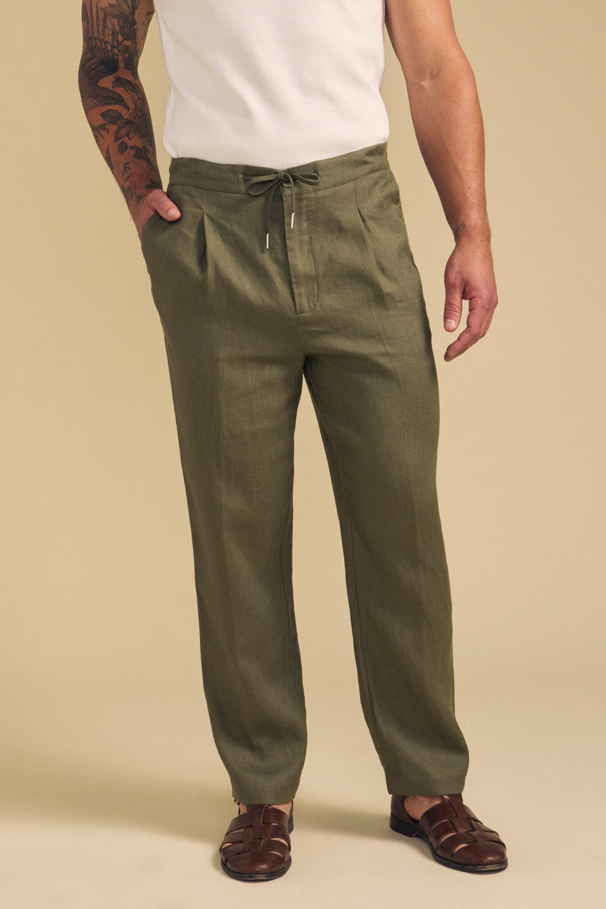 A man wearing Velvet by Graham & Spencer's ELLIOTT LINEN PANT in olive green, paired with a white sleeveless shirt and brown woven sandals, stands against a beige background.-41701921489089