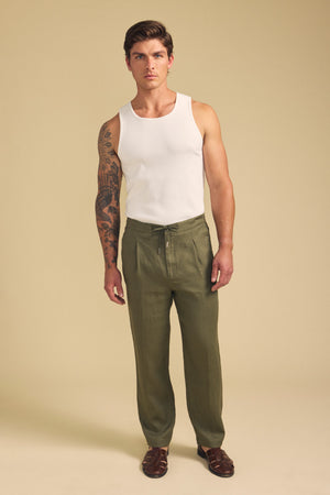 A man faces forward in a white tank top, Velvet by Graham & Spencer ELLIOTT LINEN PANT with a drawstring waist, and brown sandals against a beige background. One arm has a tattoo sleeve.