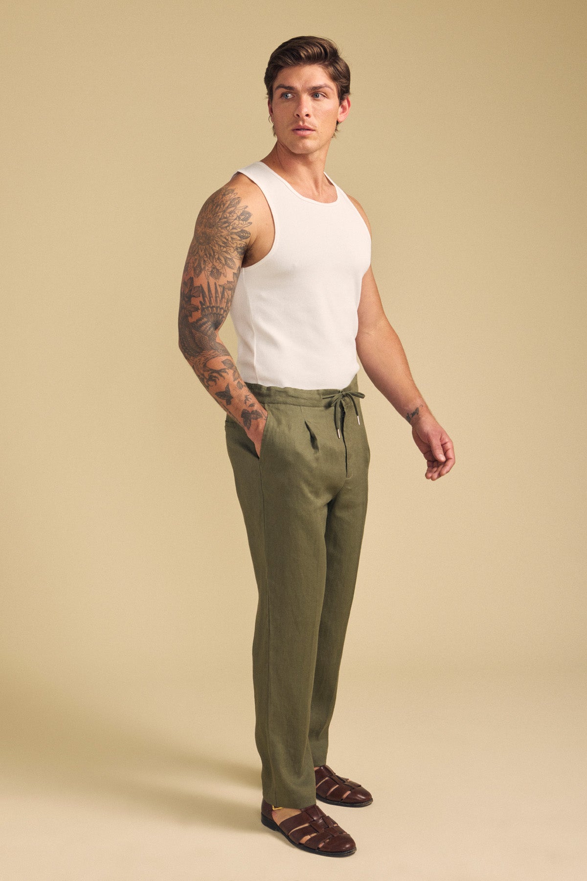 A man wearing a white tank top, Velvet by Graham & Spencer ELLIOTT LINEN PANT in olive green, and brown sandals stands against a beige background.-41701921325249