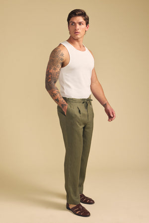 A man wearing a white tank top, Velvet by Graham & Spencer ELLIOTT LINEN PANT in olive green, and brown sandals stands against a beige background.
