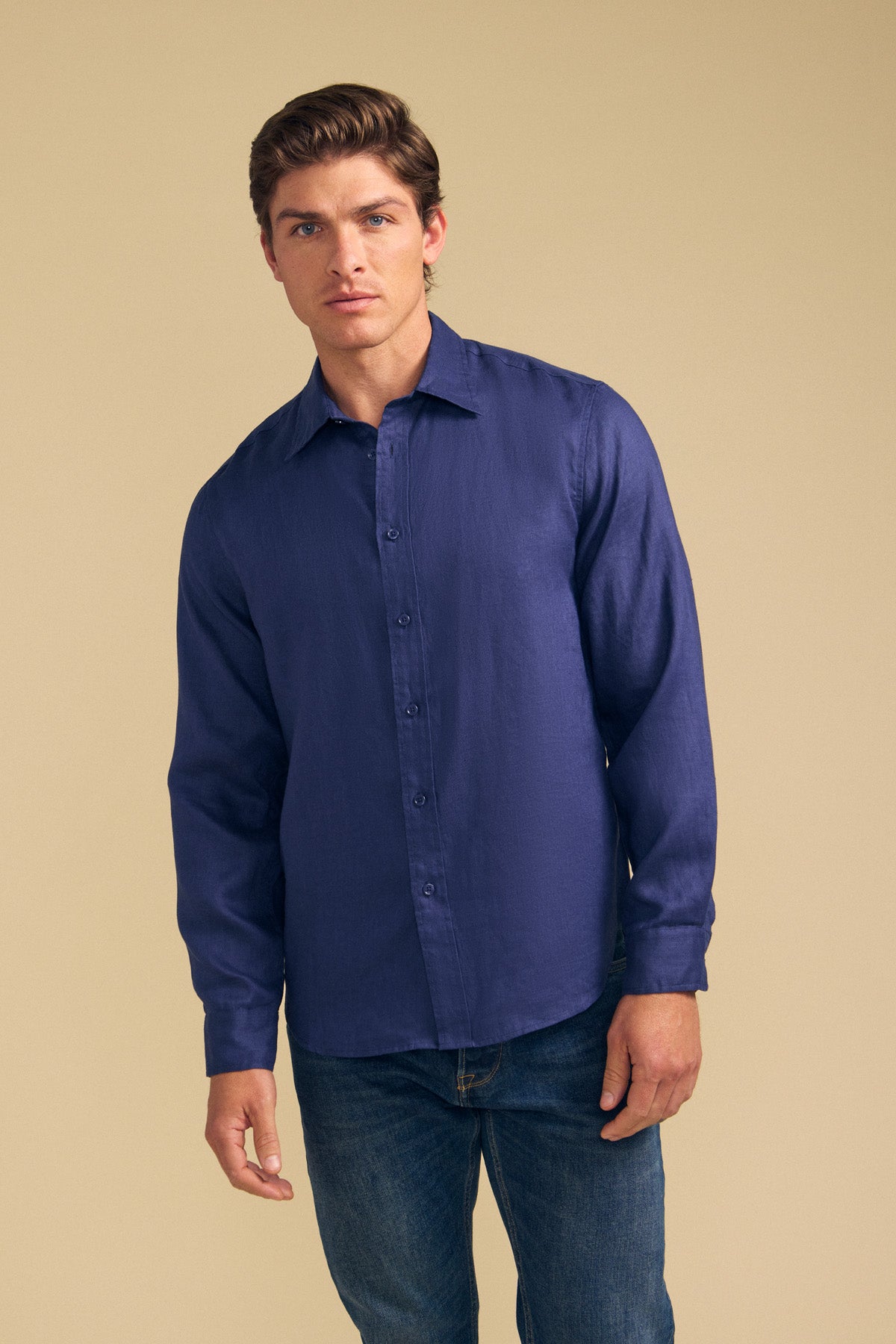 A man stands before a beige background wearing the Velvet by Graham & Spencer CARUSO LINEN SHIRT, a long-sleeve dark blue button-up, paired with blue jeans to showcase its versatile style.