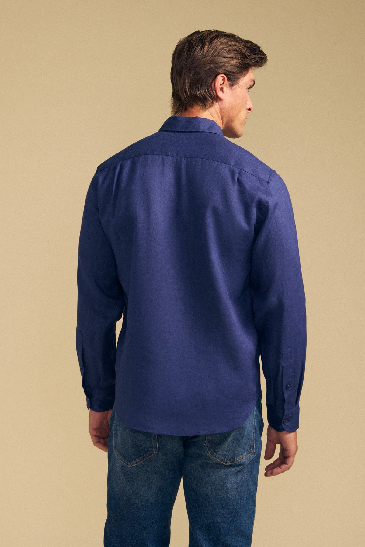 A man stands facing away from the camera against a beige background, wearing the CARUSO LINEN SHIRT by Velvet by Graham & Spencer with blue jeans—showcasing its versatile styling that's perfect for effortless layering.-41701813387457