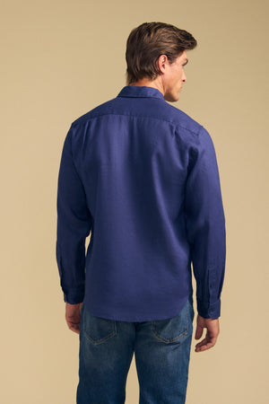 A man stands facing away from the camera against a beige background, wearing the CARUSO LINEN SHIRT by Velvet by Graham & Spencer with blue jeans—showcasing its versatile styling that's perfect for effortless layering.