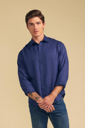 A man with short brown hair models the CARUSO LINEN SHIRT by Velvet by Graham & Spencer—its blue button-up style paired with jeans against a beige backdrop makes it perfect for versatile styling and effortless layering.
