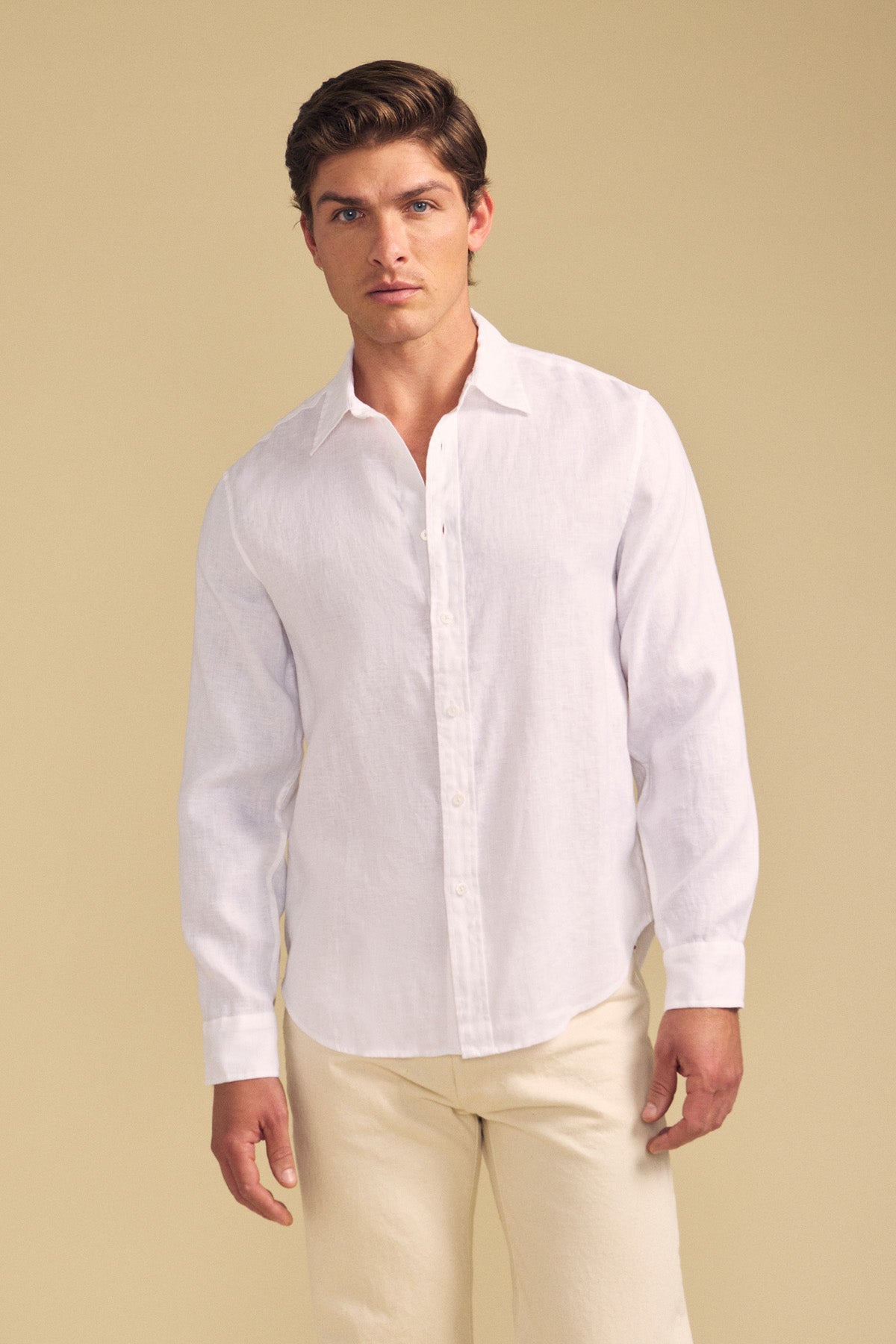 A man models the CARUSO LINEN SHIRT by Velvet by Graham & Spencer—its crisp white, long-sleeve design against a beige backdrop highlights versatile style fit for any occasion.-41701813223617