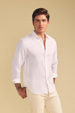 A man stands against a beige background wearing the CARUSO LINEN SHIRT by Velvet by Graham & Spencer, a white button-up long-sleeve ideal for versatile styling and effortless layering.