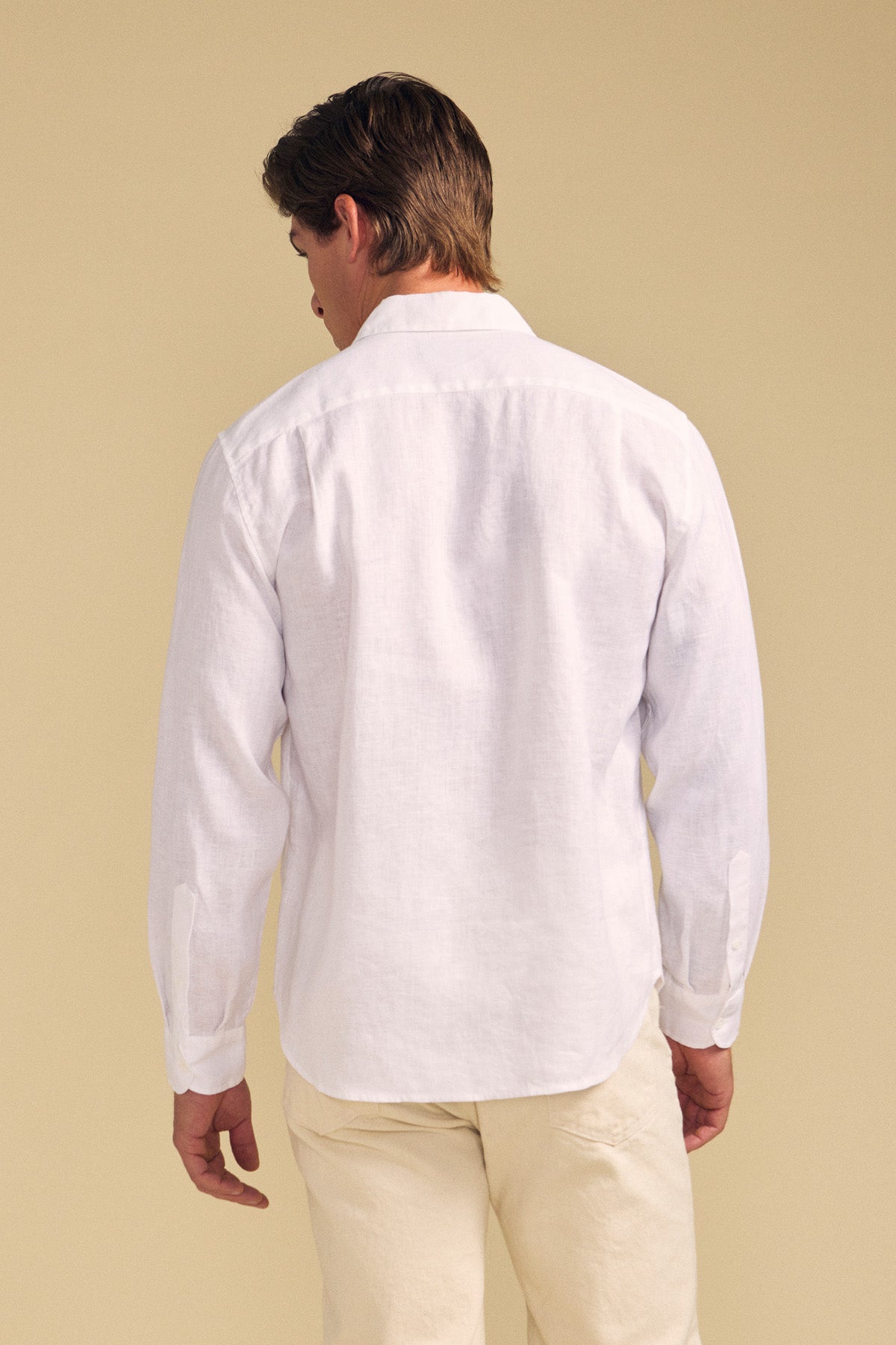 A person stands facing away, wearing the CARUSO LINEN SHIRT by Velvet by Graham & Spencer with light-colored pants against a beige background—ideal for versatile styling.-41701813027009