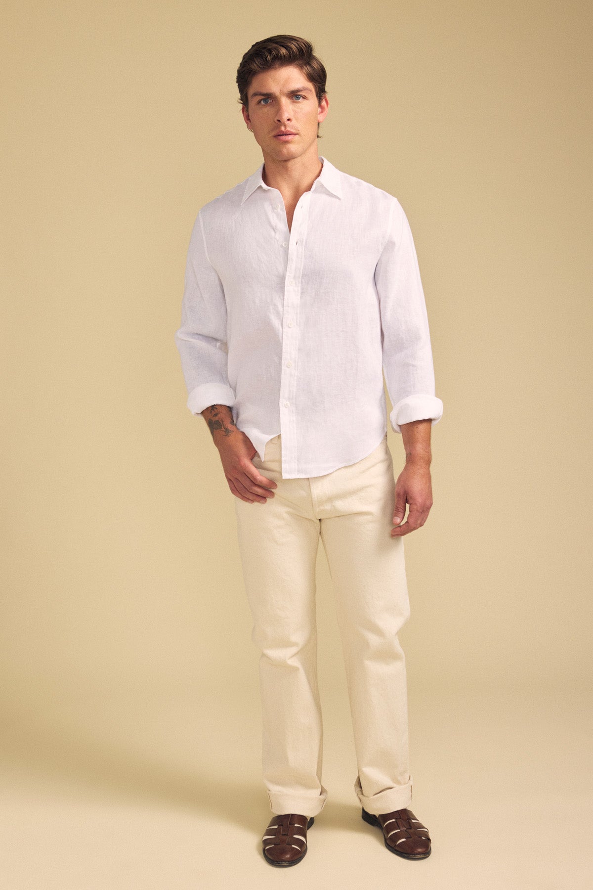 A man stands against a beige background wearing the CARUSO LINEN SHIRT by Velvet by Graham & Spencer, light beige pants, and brown sandals—demonstrating versatile styling and effortless layering for any occasion.
