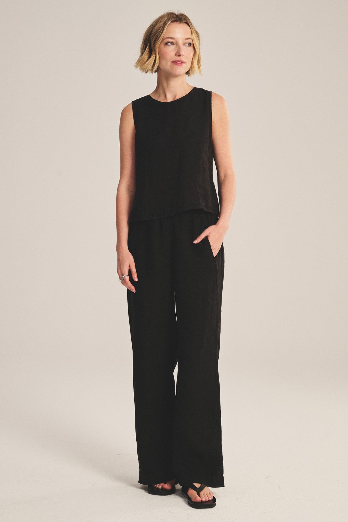 A woman stands against a plain light background, wearing Velvet by Graham & Spencer's LOLA LINEN PANT—lightweight linen trousers with an elastic waist—paired with a sleeveless black top and one hand in her pocket.