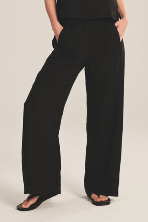 A person models the Velvet by Graham & Spencer LOLA LINEN PANT—loose-fitting, black pull-on pants with an elastic waist—paired with black sandals, hands in pockets, standing against a plain background.