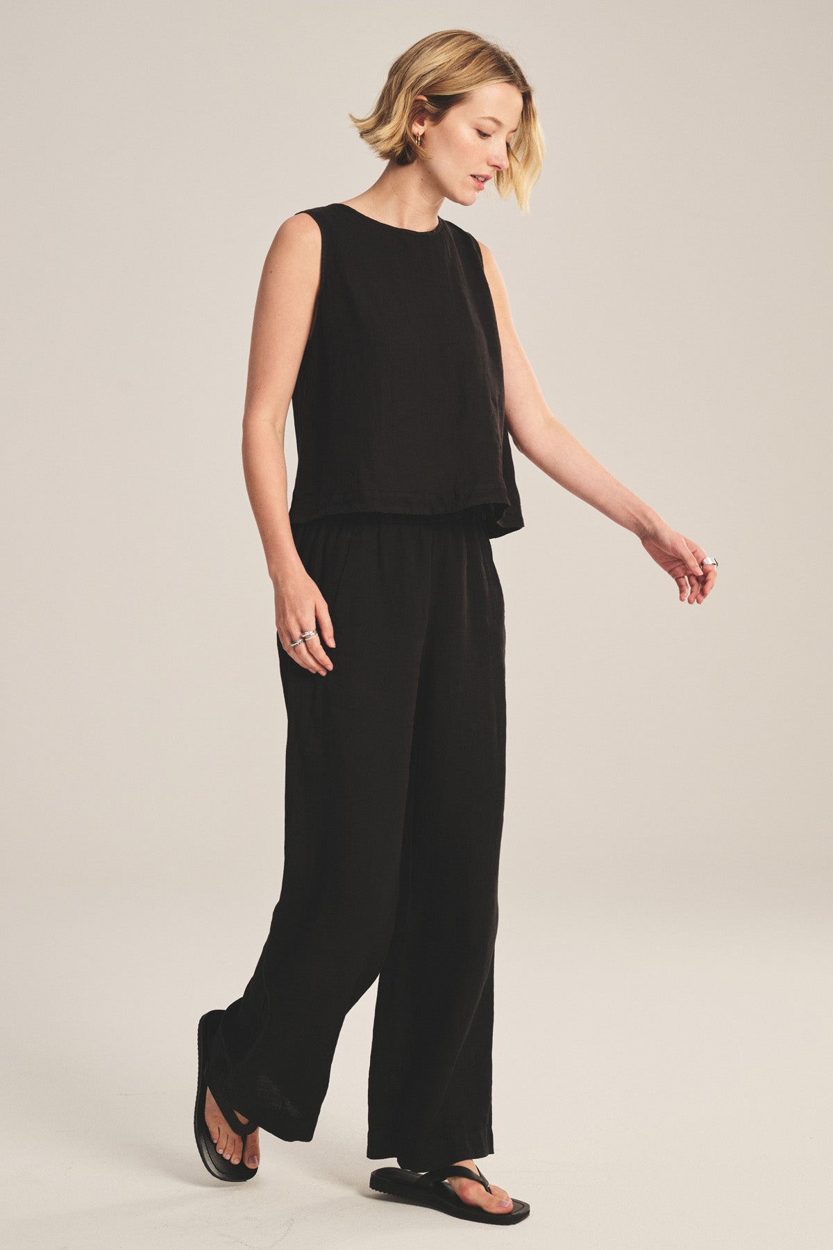 A person stands looking to the side, wearing a sleeveless black top, Velvet by Graham & Spencer LOLA LINEN PANT with an elastic waist, black sandals, and hoop earrings against a plain light background.