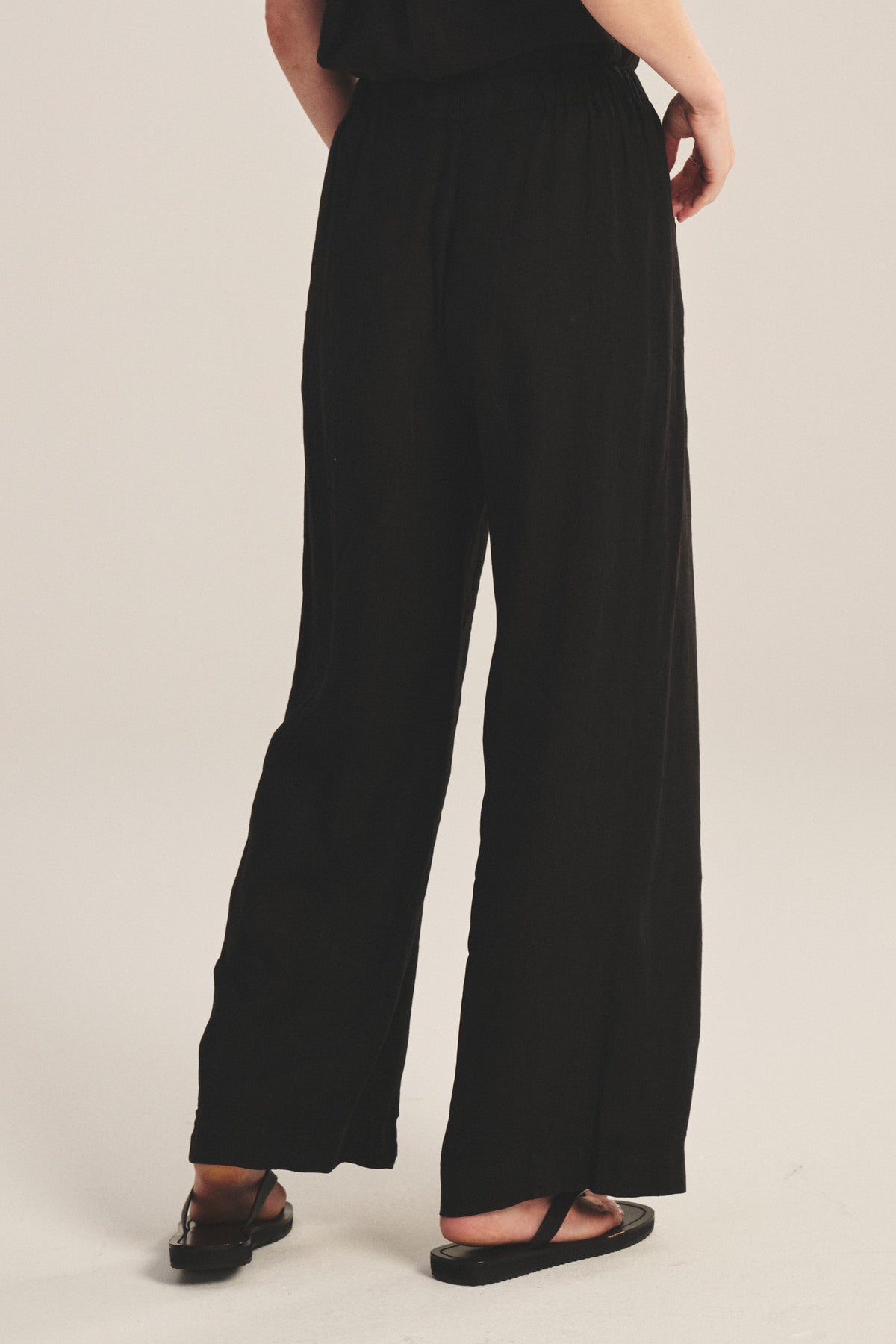 A person is shown from the back wearing the Velvet by Graham & Spencer LOLA LINEN PANT—loose-fitting, black, lightweight linen trousers with an elastic waist—paired with black sandals on a light background.