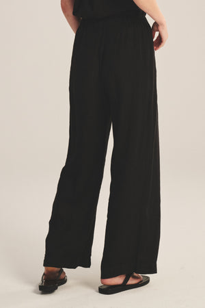 A person is shown from the back wearing the Velvet by Graham & Spencer LOLA LINEN PANT—loose-fitting, black, lightweight linen trousers with an elastic waist—paired with black sandals on a light background.