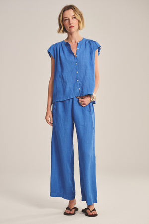 A woman stands against a plain background wearing Velvet by Graham & Spencer’s LOLA LINEN PANT with an elastic waist, paired with a loose-fitting blue short-sleeve button-up top and black sandals.