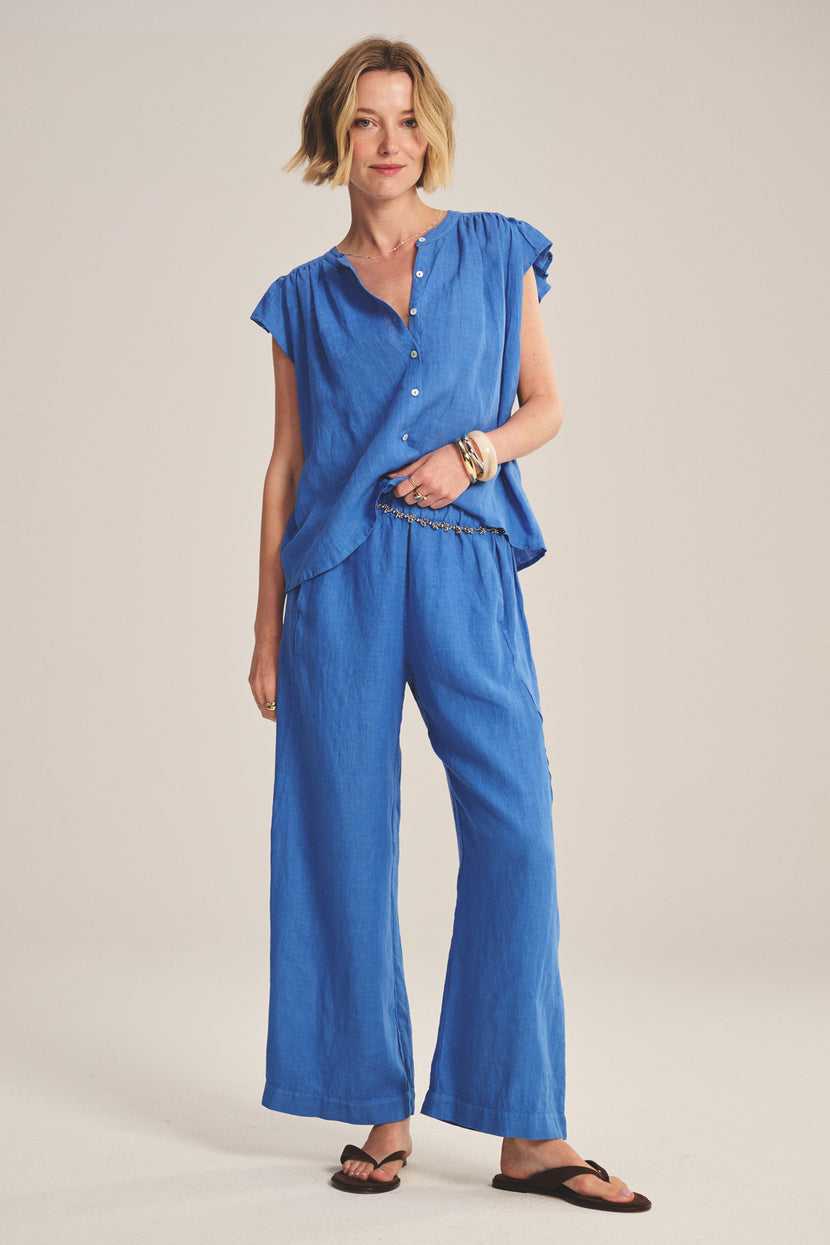 A woman stands smiling in Velvet by Graham & Spencer’s LOLA LINEN PANT, a lightweight pull-on trouser with an elastic waist, paired with a loose blue short-sleeve buttoned top, bracelets, and black sandals against a plain background.