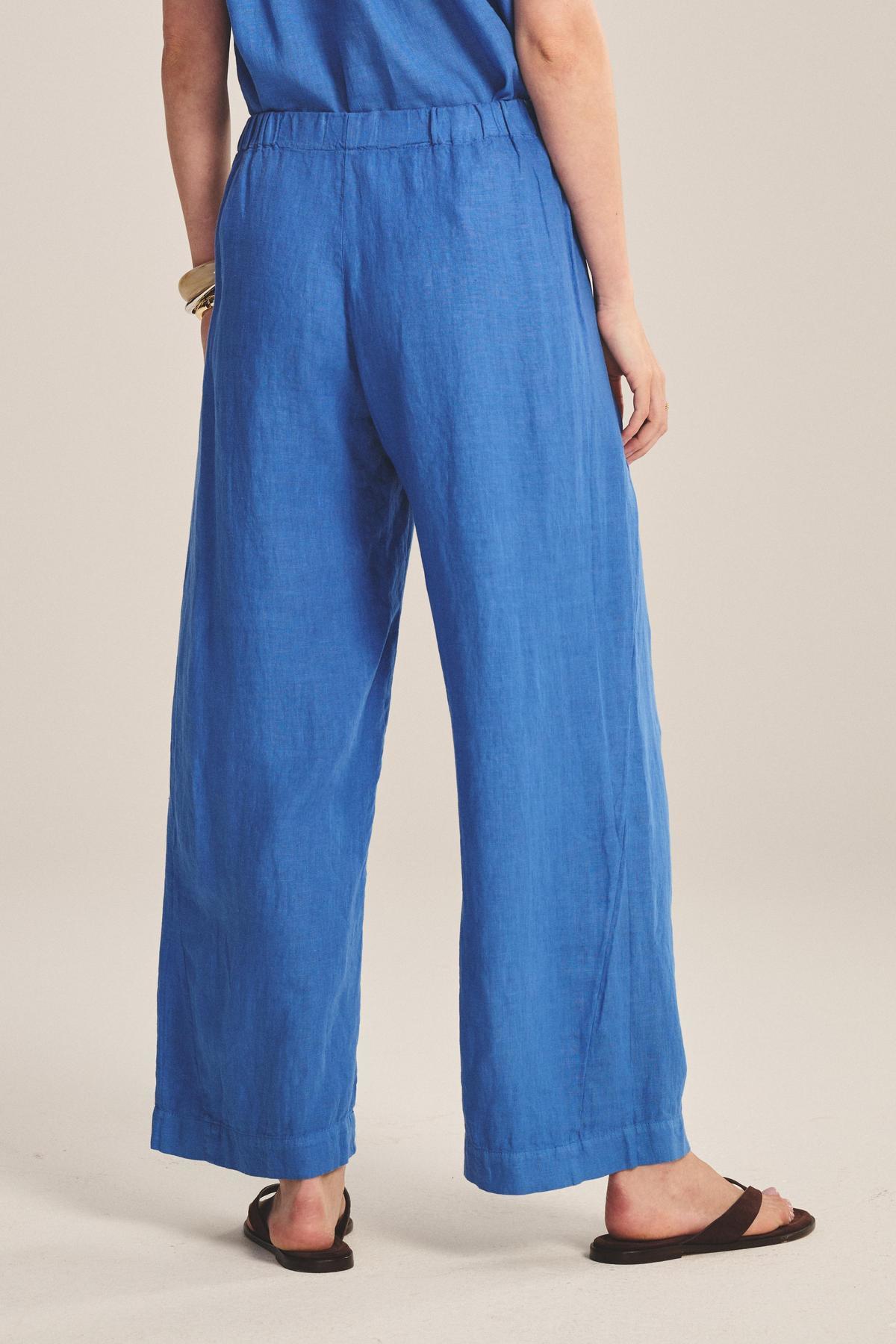 Person stands with back to camera in Velvet by Graham & Spencer's LOLA LINEN PANT, featuring a loose fit and elastic waist, paired with a blue top, black sandals, and gold bracelets against a plain background.
