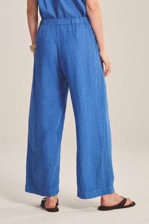 Person stands with back to camera in Velvet by Graham & Spencer's LOLA LINEN PANT, featuring a loose fit and elastic waist, paired with a blue top, black sandals, and gold bracelets against a plain background.