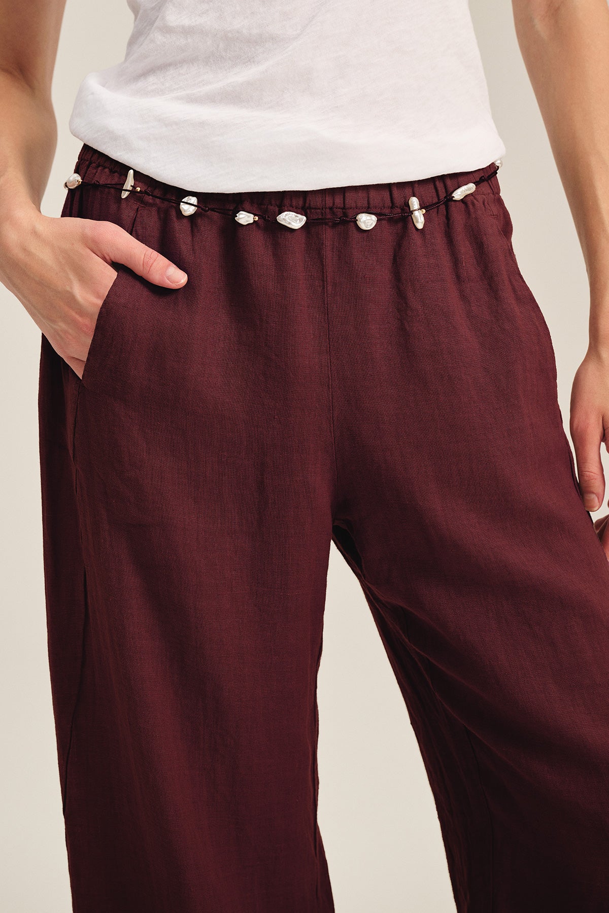   A person wears Velvet by Graham & Spencer's LOLA LINEN PANT in burgundy, styled with a white shirt. The pull-on pants feature an elastic waist, a drawstring with white seashells, and have one hand tucked into the pocket. 