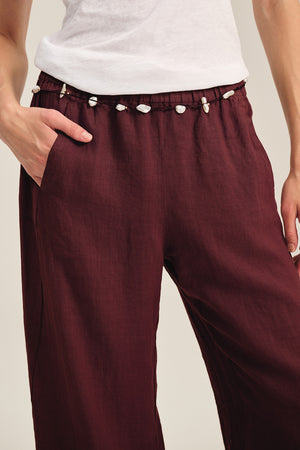 A person wears Velvet by Graham & Spencer's LOLA LINEN PANT in burgundy, styled with a white shirt. The pull-on pants feature an elastic waist, a drawstring with white seashells, and have one hand tucked into the pocket.