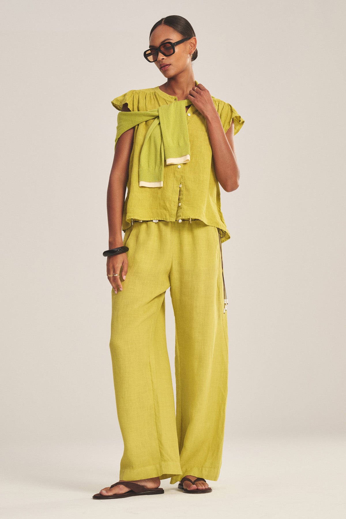 A woman in Velvet by Graham & Spencer's LOLA LINEN PANT, styled with a loose light green top, sweater over her shoulder, dark sandals, black sunglasses, and accessories, stands against a plain background.-41128091353281