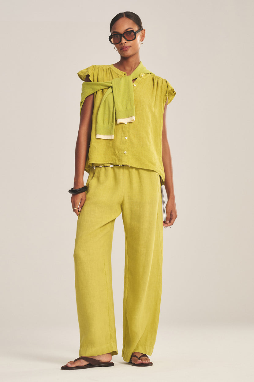 A woman stands against a plain background wearing Velvet by Graham & Spencer’s LOLA LINEN PANT in light green—a relaxed, straight-leg pull-on style—with a matching button-up top, draped sweater, sunglasses, and black sandals.