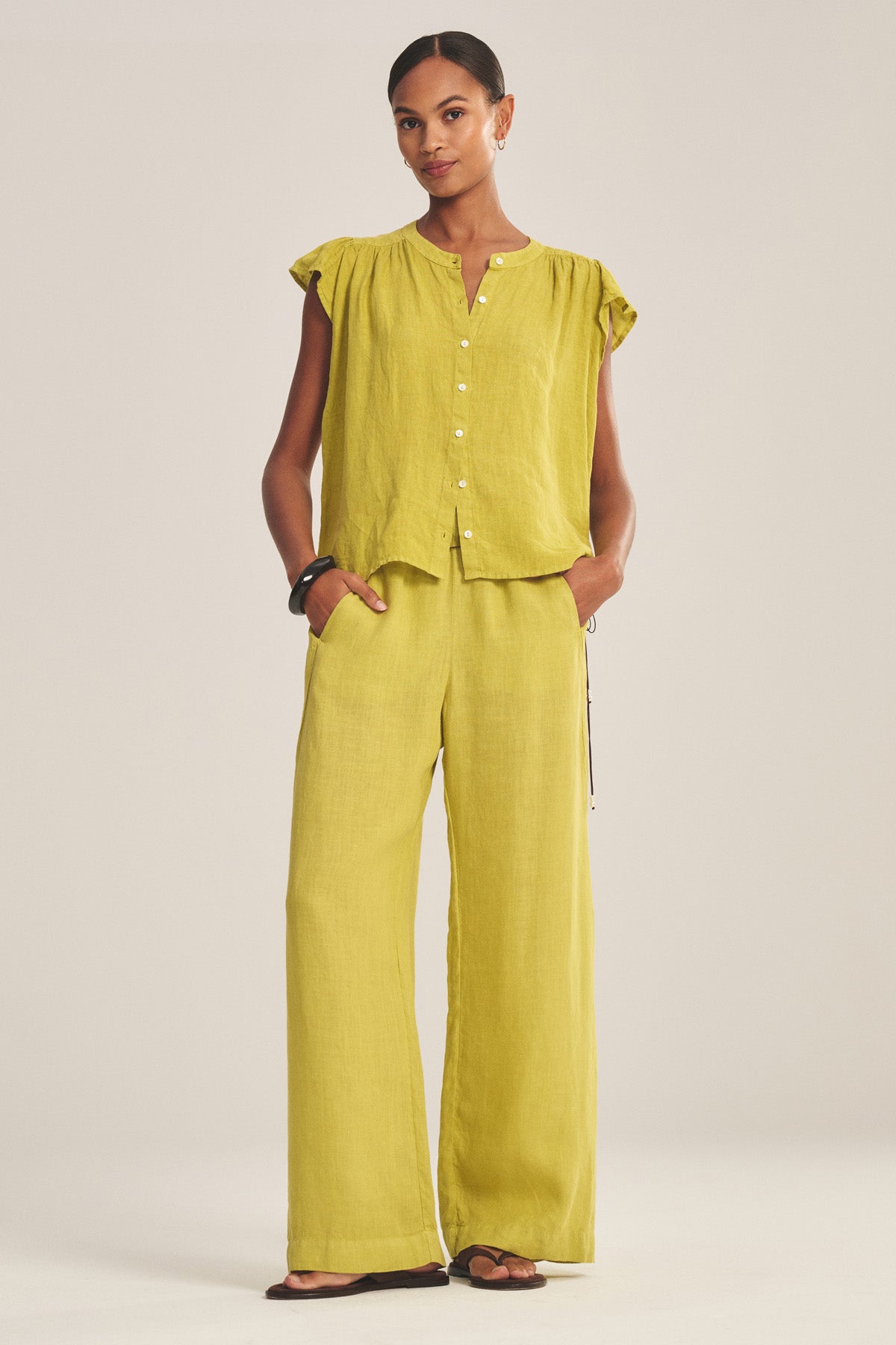 A woman stands facing forward, wearing Velvet by Graham & Spencer's LOLA LINEN PANT and a matching loose-fitting, short-sleeve, button-up yellow top with sandals. Her hands are in her pockets.-41128091287745