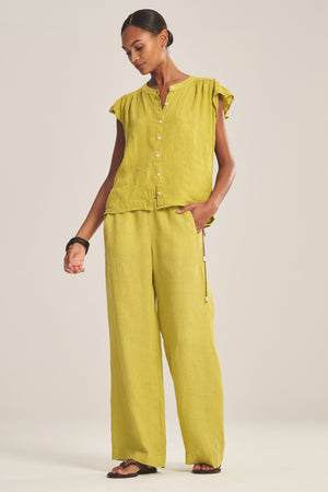A woman stands against a plain background wearing the LOLA LINEN PANT by Velvet by Graham & Spencer—a pair of relaxed, straight-leg, lightweight linen pull-on pants in matching light green—styled with a loose-fitting, ruffled-sleeve button-up top, black sandals, and black bracelets.