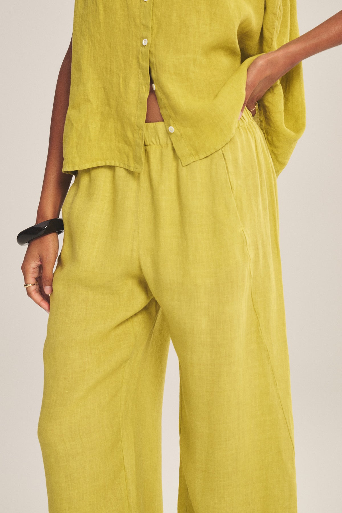 A person wears the LOLA LINEN PANT by Velvet by Graham & Spencer—relaxed, straight-leg, pull-on pants in yellow-green—paired with a matching loose linen shirt, one hand in a pocket and a black bangle on their wrist.-41128091222209