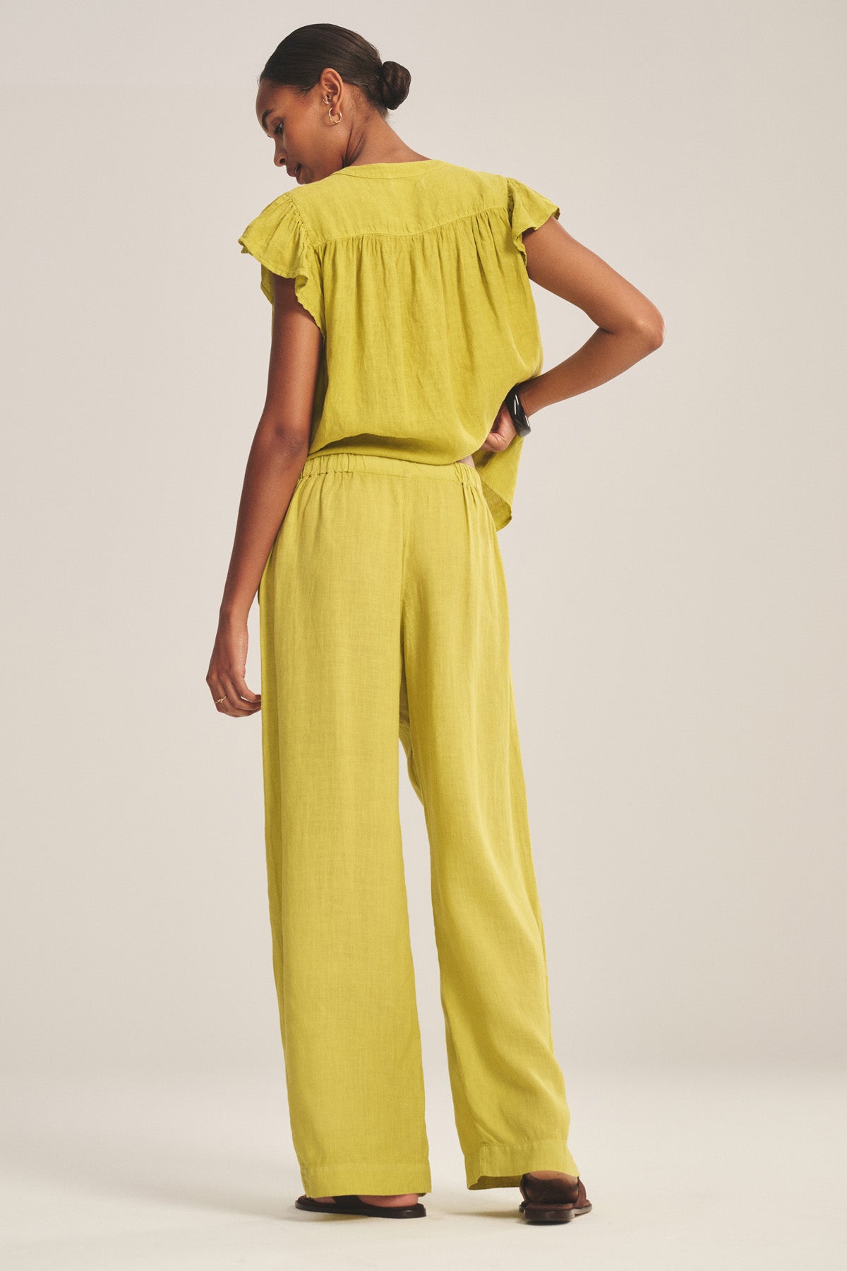 A woman faces away, wearing the Velvet by Graham & Spencer LOLA LINEN PANT—lightweight linen with a relaxed straight leg—and a matching loose yellow top and sandals against a plain background.-41128091156673