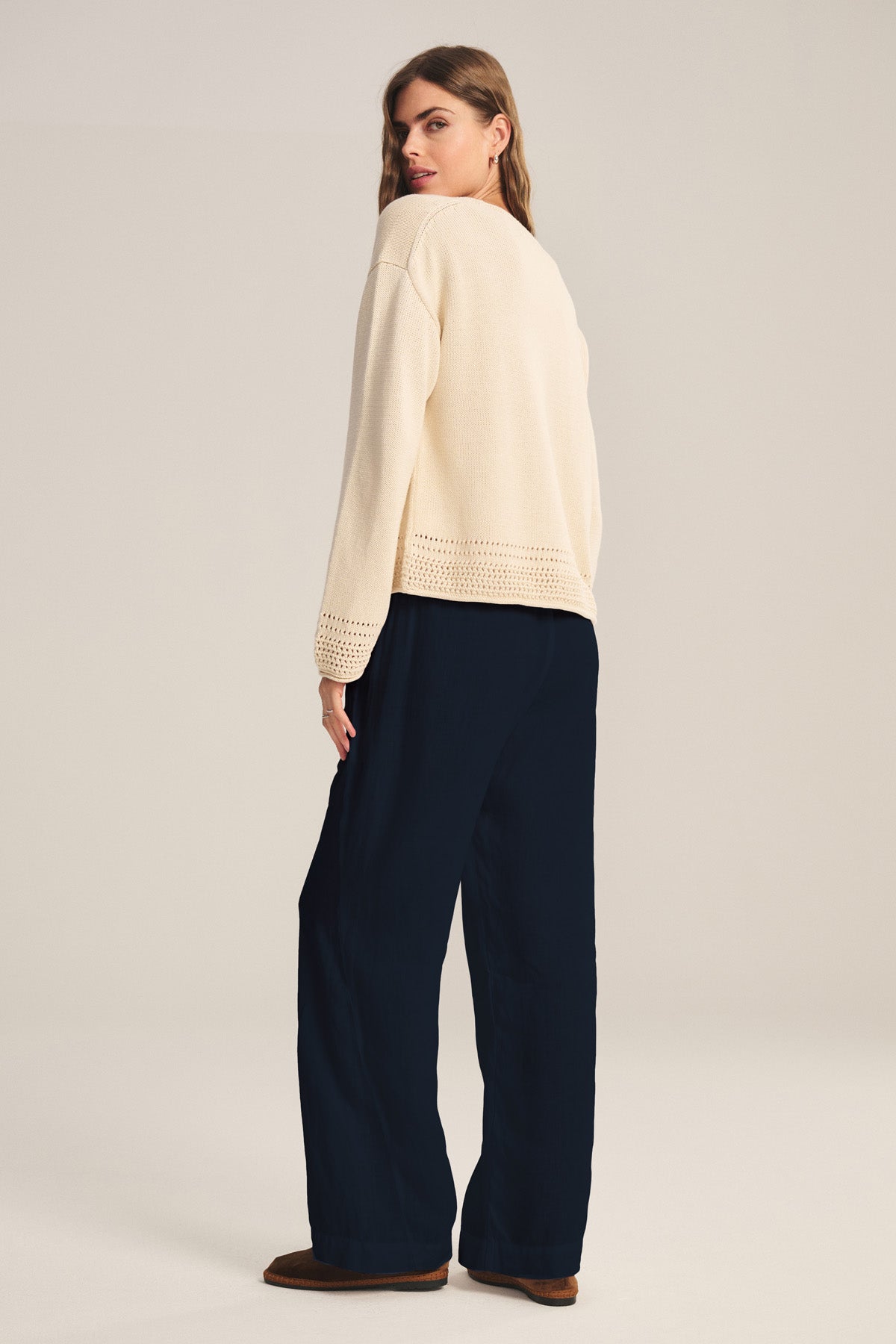 A woman stands facing slightly backward, wearing a cream long-sleeve sweater with cutout hem, Velvet by Graham & Spencer LOLA LINEN PANT in navy with an elastic waist, and brown shoes against a neutral background.