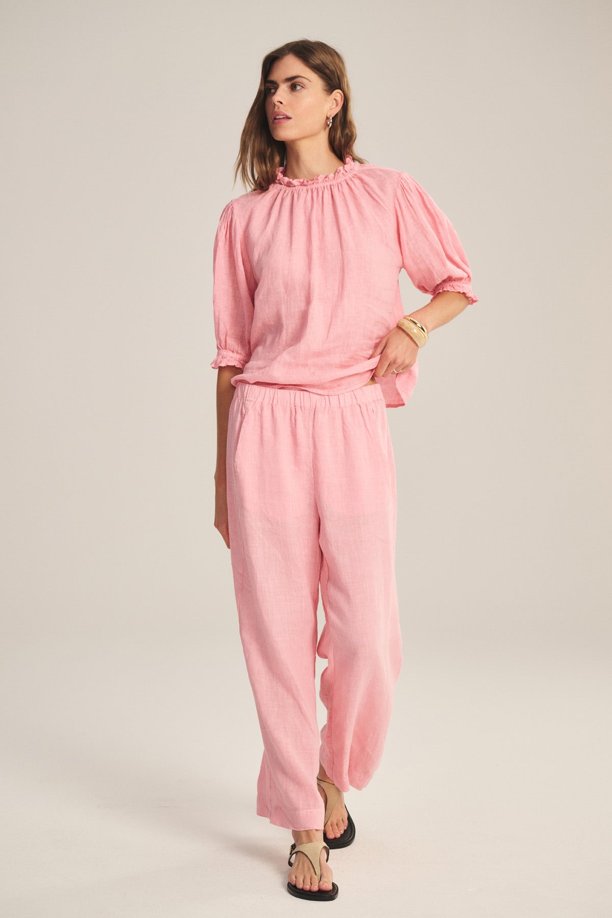 A person wears the Velvet by Graham & Spencer LOLA LINEN PANT, a lightweight, elastic-waist linen pant in pink, paired with a matching blouse, black sandals, and gold bracelets against a simple light background.-41296714989761