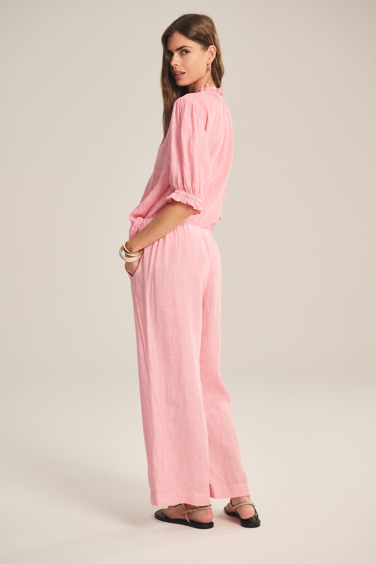 A person stands sideways against a plain background, wearing Velvet by Graham & Spencer's LOLA LINEN PANT—a light pink, loose-fitting, lightweight linen pant with an elastic waistband—styled with a coordinating top and black flats.-41296714956993