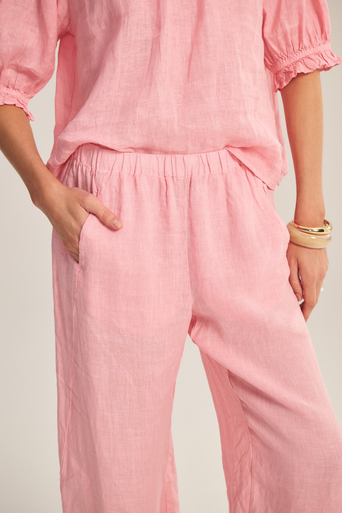 A person wearing the LOLA LINEN PANT by Velvet by Graham & Spencer—a pair of relaxed, straight-leg pull-on pants in light pink linen with a loose fit—matches their short-sleeve top, left hand in pocket and gold bangles adorning their right wrist.