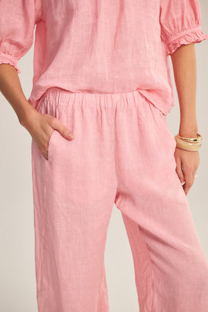 A person wearing the LOLA LINEN PANT by Velvet by Graham & Spencer—a pair of relaxed, straight-leg pull-on pants in light pink linen with a loose fit—matches their short-sleeve top, left hand in pocket and gold bangles adorning their right wrist.