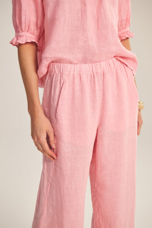 Person wearing the LOLA LINEN PANT by Velvet by Graham & Spencer—relaxed, straight-leg linen pants with an elastic waistband, paired with a loose-fitting, light pink linen blouse, one hand in the pocket.