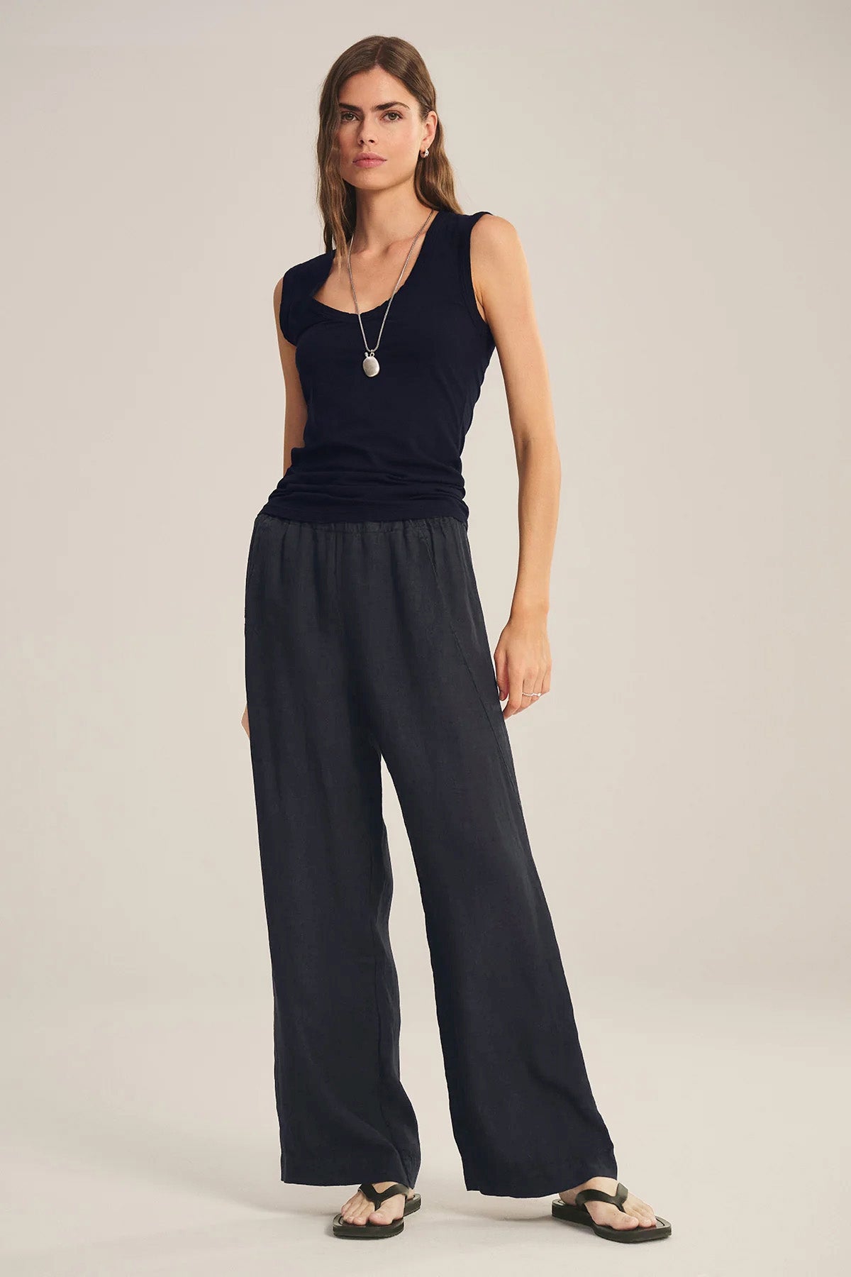 A woman wears the LOLA LINEN PANT by Velvet by Graham & Spencer with a black sleeveless top, black sandals, and a long necklace against a plain background.-41073343987905
