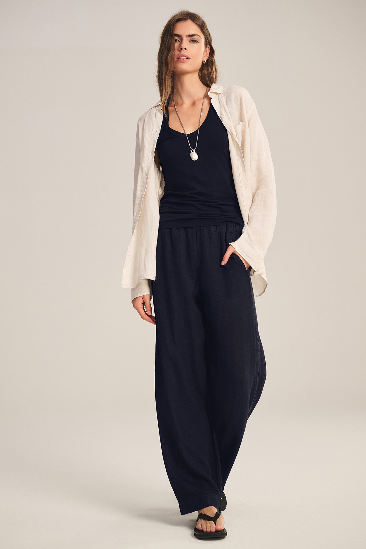   A person stands against a neutral background wearing Velvet by Graham & Spencer's LOLA LINEN PANT, a cream button-up shirt over a black top, black sandals, and a necklace. 