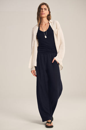 A person stands against a neutral background wearing Velvet by Graham & Spencer's LOLA LINEN PANT, a cream button-up shirt over a black top, black sandals, and a necklace.