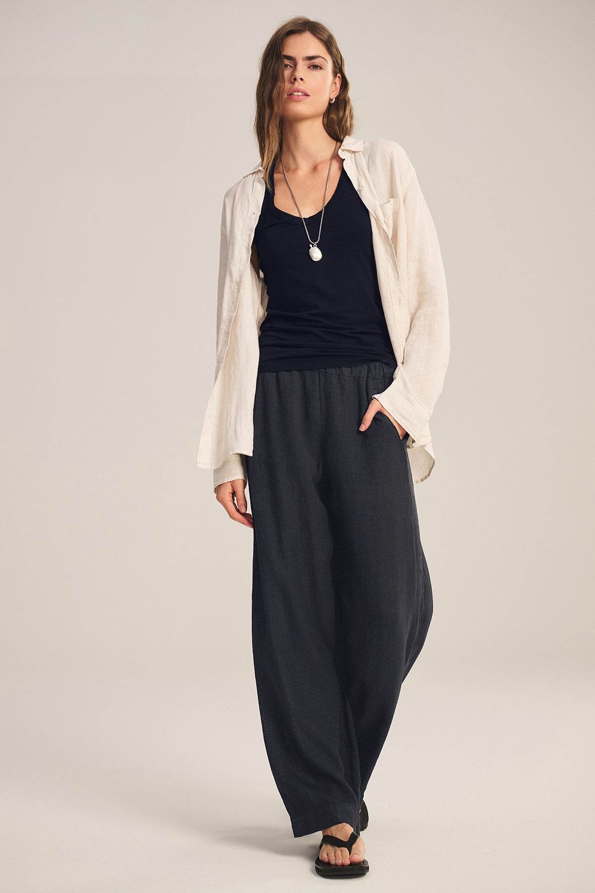 A person stands in a neutral studio wearing the LOLA LINEN PANT by Velvet by Graham & Spencer, styled with a cream shirt over a black top, black sandals, and a necklace.-41081093488833