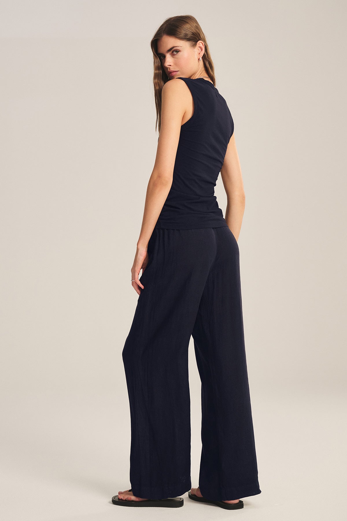 A woman stands facing slightly away, wearing a sleeveless black top, black sandals, and Velvet by Graham & Spencer's LOLA LINEN PANT—lightweight linen trousers with an elastic waist—set against a plain light background.-40010225811649