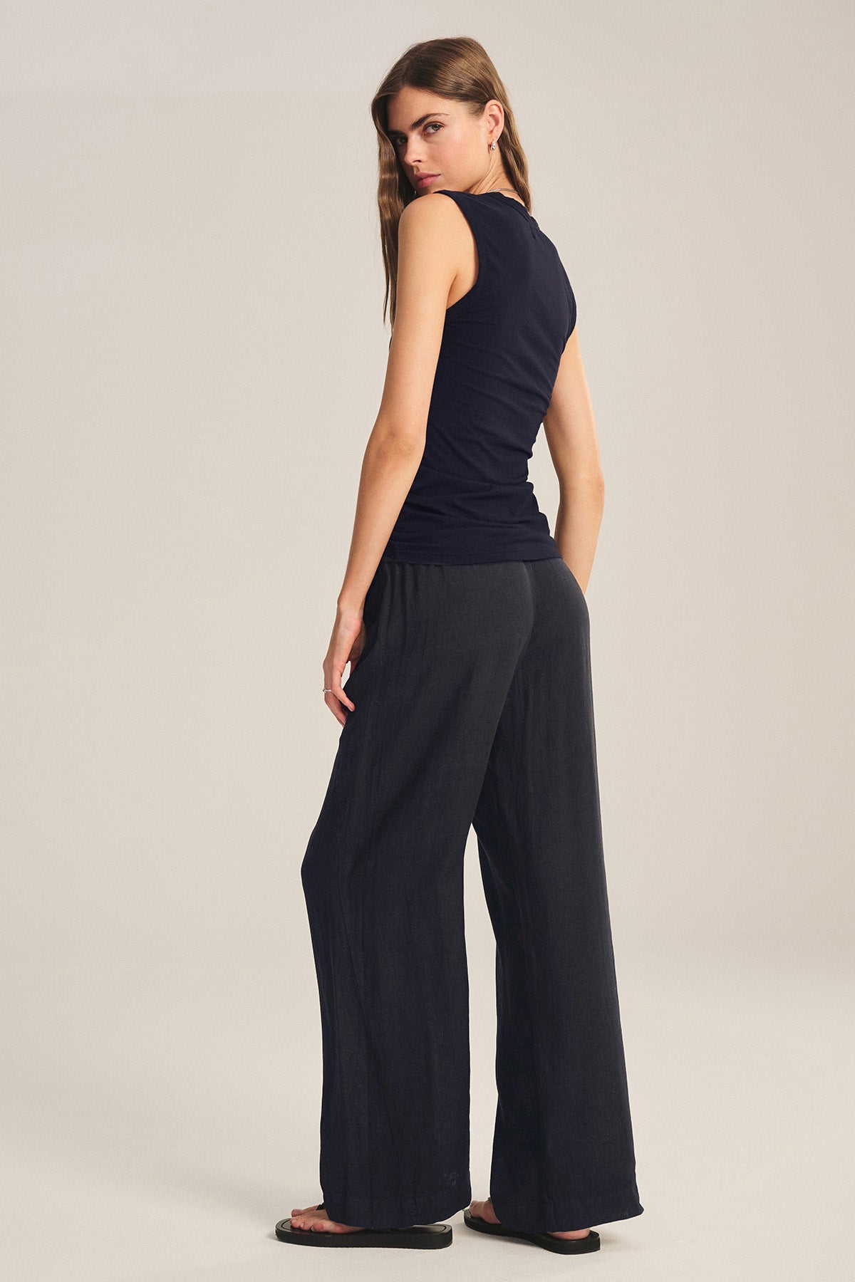 A woman faces away against a plain light background, wearing a black sleeveless top, flip-flops, and the LOLA LINEN PANT by Velvet by Graham & Spencer—lightweight trousers with an elastic waist.