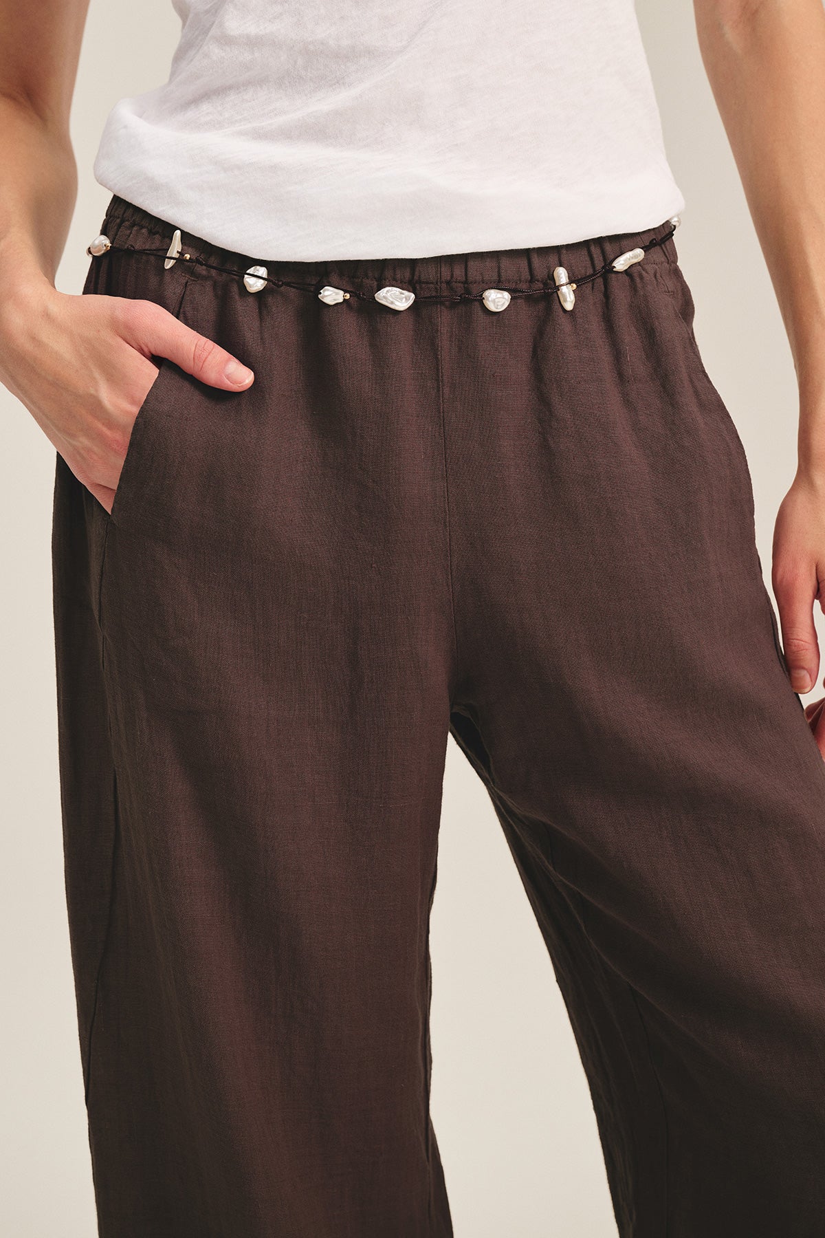   A person wears Velvet by Graham & Spencer’s LOLA LINEN PANT—a brown, lightweight linen trouser with an elastic waist adorned with small white shells—paired with a white shirt, one hand in the pocket. 