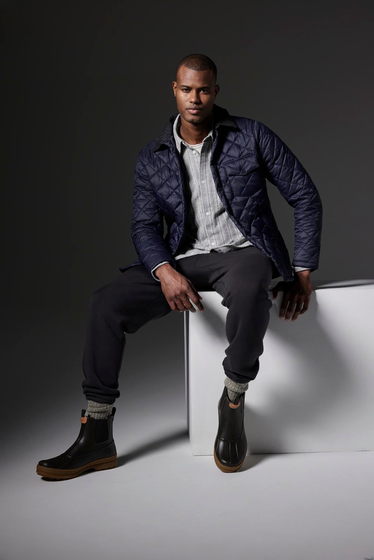   In a plain dark gray studio, a man sits on a white cube wearing the LOU JACKET by Velvet by Graham & Spencer, which is a black quilted puffer jacket with a corduroy collar. He pairs it with a striped shirt, black pants, and black boots. 