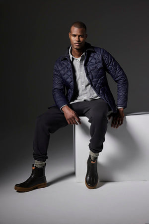 In a plain dark gray studio, a man sits on a white cube wearing the LOU JACKET by Velvet by Graham & Spencer, which is a black quilted puffer jacket with a corduroy collar. He pairs it with a striped shirt, black pants, and black boots.