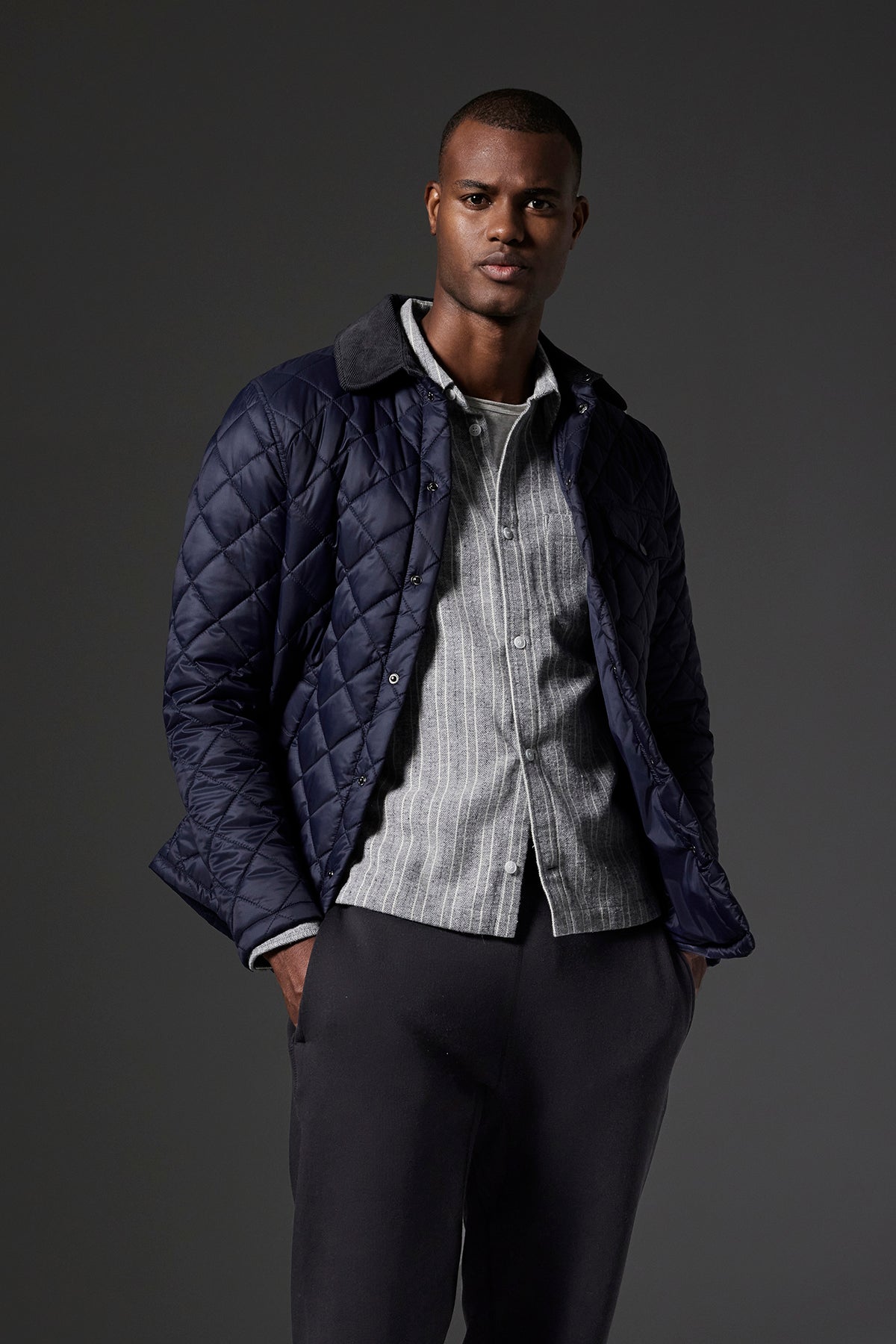   A man wearing the LOU JACKET, a dark quilted puffer jacket with a corduroy collar by Velvet by Graham & Spencer, over a light-colored shirt and dark pants stands against a dark background, with one hand in his pocket. 